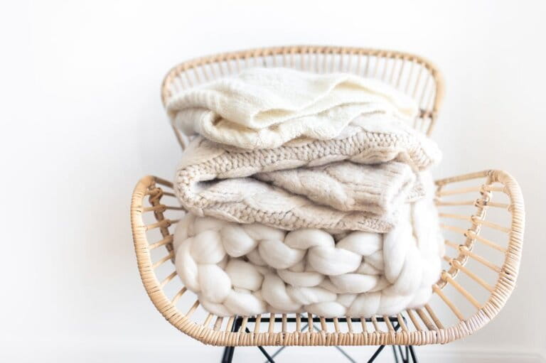 Stack of cozy wool white knitted sweaters on wicker armchair. Pile of merino winter clothes. 