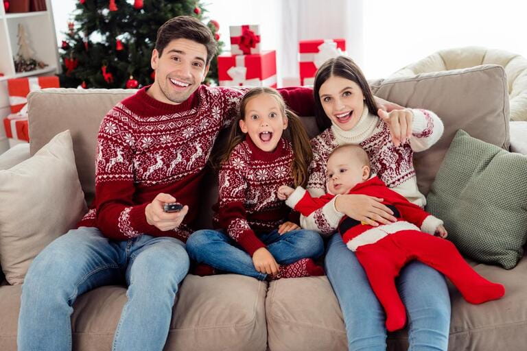Photo of impressed cute couple two kids dressed new year pullovers sitting couch watching tv pointing finger you indoors house home room