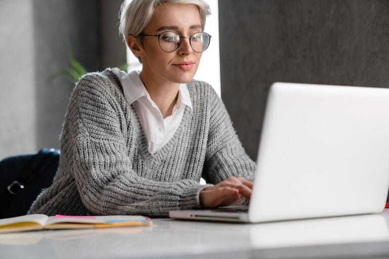 White-haired focused woman working with laptop while sitting at table in office White-haired focused woman working with laptop while sitting at table in office