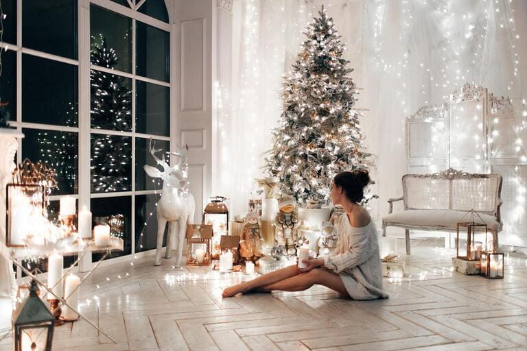 woman in white sweater, warm and cozy evening in Christmas interior design, Christmas tree decorated by light toys gifts, candles, lanterns, light garland in living room. woman in white sweater, warm and cozy evening in Christmas interior design, Christmas tree decorated by light toys gifts, candles, lanterns, light garland in living room.