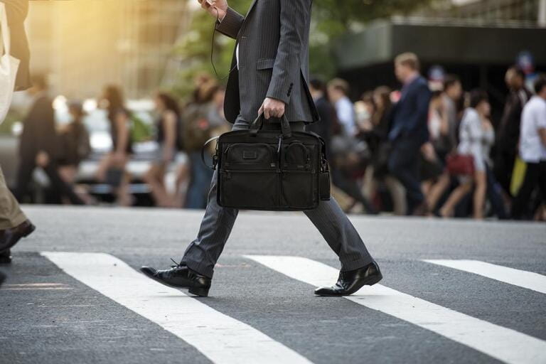 Businessman crossing the street on the footpath and holding a laptop bag and smatphone in the city