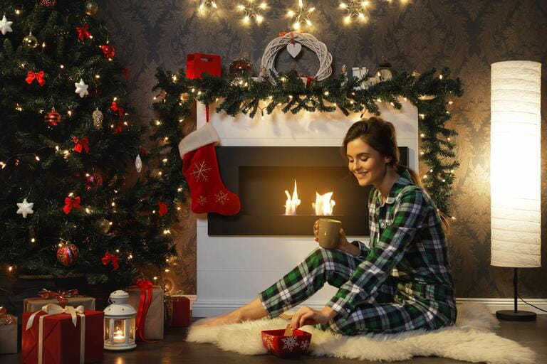 Christmas or New Year celebration. Woman sitting beside fireplace is drinking hot tea with cookies.