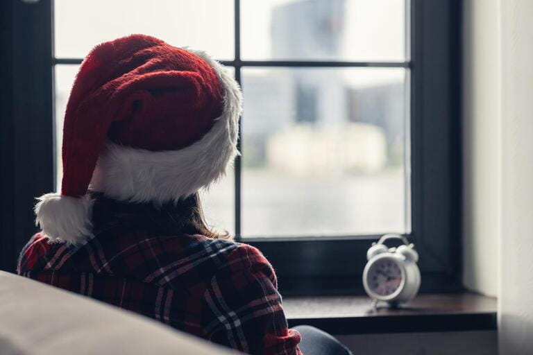 Back view of lonely sad young woman in a red santa claus christmas hat sitting near window and holding alarm clock.  Back view of lonely sad young woman in a red santa claus christmas hat sitting near window and holding alarm clock.