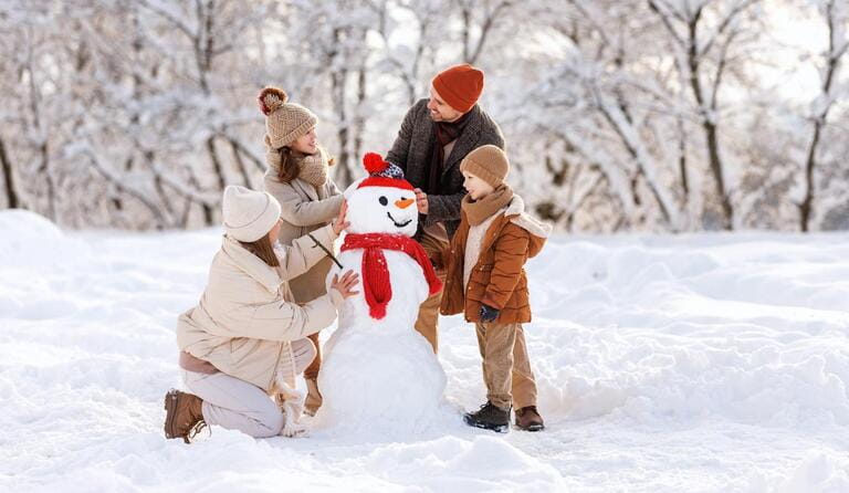 Happy parents and children gathering in snow-covered park together sculpting funny snowman from snow. Father, mother and two kids playing outdoor in winter forest. Family active holiday Happy parents and children gathering in snow-covered park together sculpting funny snowman from snow. Father, mother and two kids playing outdoor in winter forest. Family active holiday