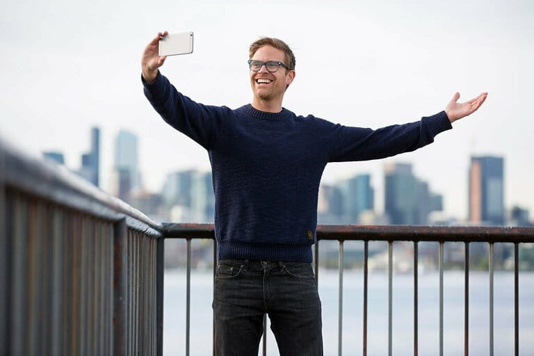 A man taking a selfie with a smart phone in font of a cityscape in the distance. A man taking a selfie with a smart phone in font of a cityscape in the distance.
